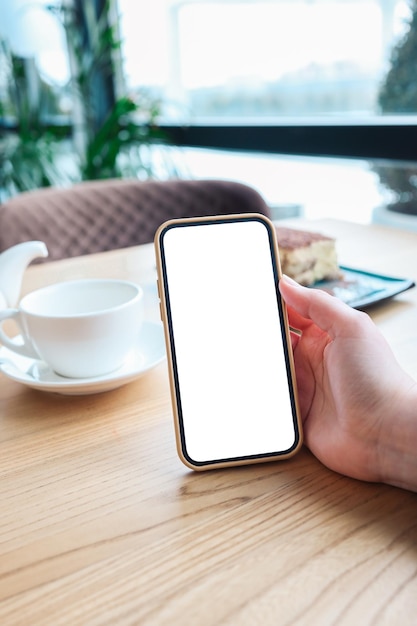 Mockup-afbeelding van een vrouw die een zwarte mobiele telefoon vasthoudt en gebruikt met een leeg desktopscherm terwijl ze thee drinkt in café