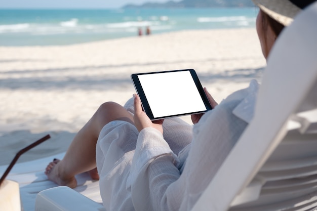 Mockup-afbeelding van een vrouw die een zwarte tablet-pc met een leeg bureaubladscherm vasthoudt terwijl ze op een strandstoel op het strand ligt