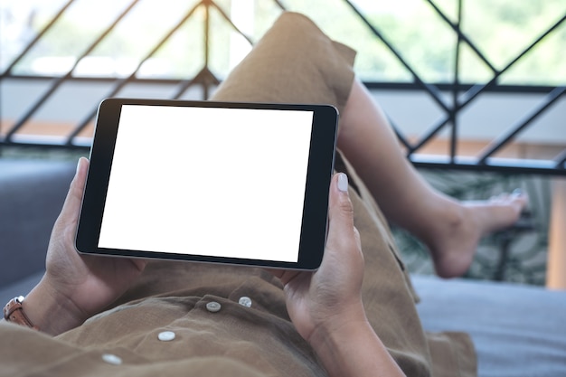 Mockup-afbeelding van een vrouw die een zwarte tablet-pc met een leeg wit bureaublad horizontaal houdt terwijl ze in de woonkamer ligt met een ontspannen gevoel
