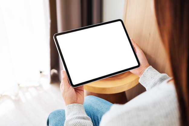 Mockup-afbeelding van een vrouw die een zwarte tablet-pc vasthoudt met een leeg wit bureaubladscherm terwijl ze thuis op een gezellig wit bed zit