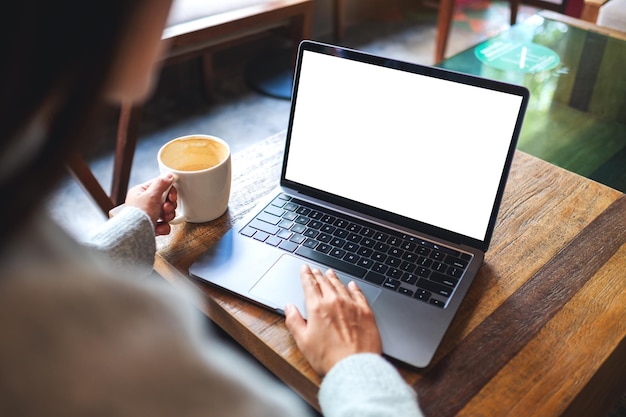 Mockup-afbeelding van een vrouw die laptop-touchpad gebruikt en aanraakt met een leeg wit bureaubladscherm terwijl ze koffie drinkt in café