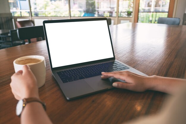 Mockup-afbeelding van een vrouw die laptop touchpad met leeg wit bureaublad op houten tafel gebruikt en aanraakt terwijl ze koffie drinkt