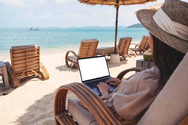 Mockup-afbeelding van een vrouw die op een laptopcomputer met een leeg bureaubladscherm typt en gebruikt terwijl ze op het strand zit