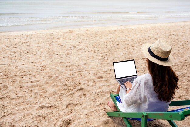 Mockup-afbeelding van een vrouw die op een laptopcomputer met een leeg desktopscherm typt en gebruikt terwijl ze op een strandstoel zit