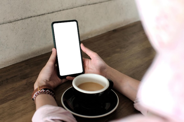 Foto mockup-afbeelding van een vrouw met een mobiele telefoon met een leeg scherm met een koffiekopje op een houten tafel