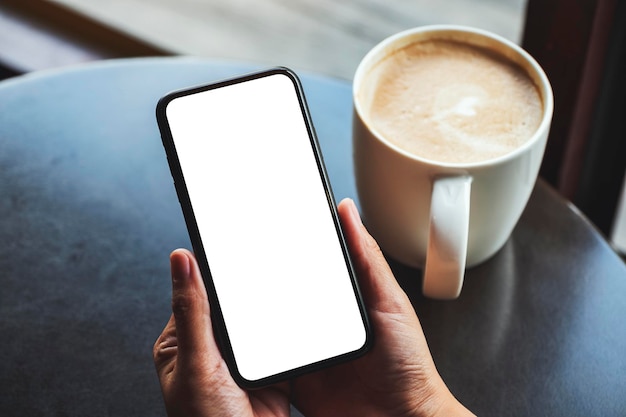 Mockup-afbeelding van een vrouw met een mobiele telefoon met een leeg wit scherm met een koffiekopje op tafel