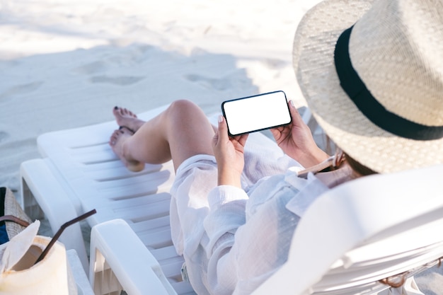 Mockup-afbeelding van een vrouw met een zwarte mobiele telefoon met een leeg desktopscherm terwijl ze op een strandstoel op het strand ligt