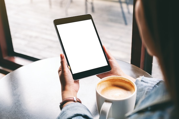 Mockup-afbeelding van een vrouw met een zwarte tablet met een wit leeg scherm en een koffiekopje op tafel