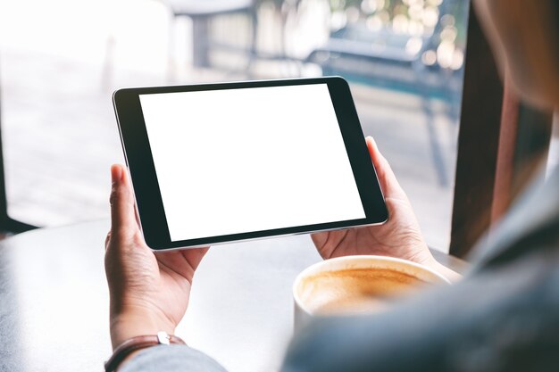 Mockup-afbeelding van een vrouw met een zwarte tablet met een wit leeg scherm en een koffiekopje op tafel