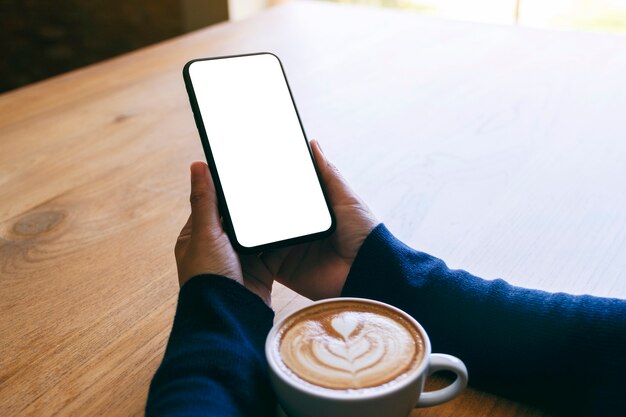 Mockup afbeelding van een vrouw met witte mobiele telefoon met leeg scherm met koffiekopje op houten tafel