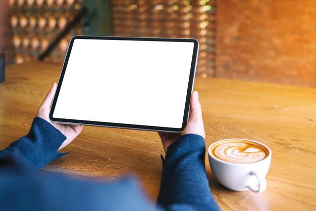 Mockup afbeelding van een vrouw met zwarte tablet pc met leeg wit scherm met koffiekopje op houten tafel