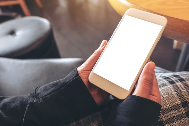 Mockup-afbeelding van handen die een witte mobiele telefoon met leeg scherm vasthouden en gebruiken terwijl u in café zit