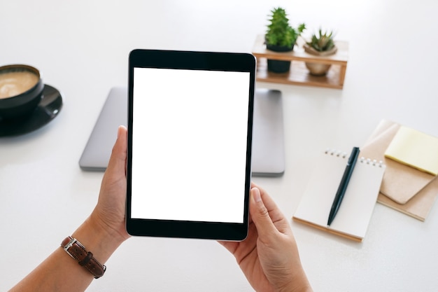 Mockup-afbeelding van handen met een tablet met een leeg wit scherm en een laptopcomputer op een houten tafel in kantoor