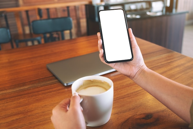 Mockup-afbeelding van handen met zwarte mobiele telefoon met leeg bureaublad tijdens het drinken van koffie in café