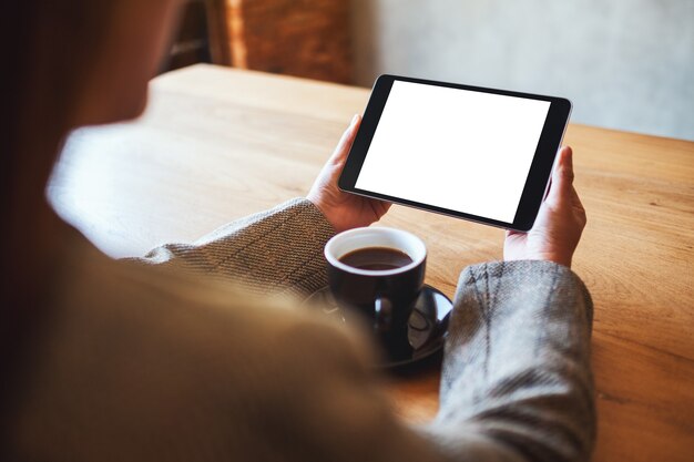 Mockup afbeelding van vrouw met zwarte tablet pc met leeg wit scherm met koffiekopje op houten tafel