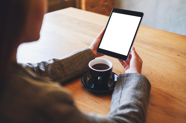 Mockup afbeelding van vrouw met zwarte tablet pc met leeg wit scherm met koffiekopje op houten tafel