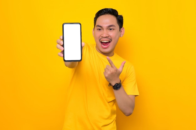 흉내낸 빈 화면 휴대폰 노란색 배경에 격리된 모바일 앱을 추천하는 빈 화면이 있는 휴대전화를 보여주는 캐주얼 티셔츠를 입은 쾌활한 젊은 아시아 남자 사람들의 라이프 스타일 개념