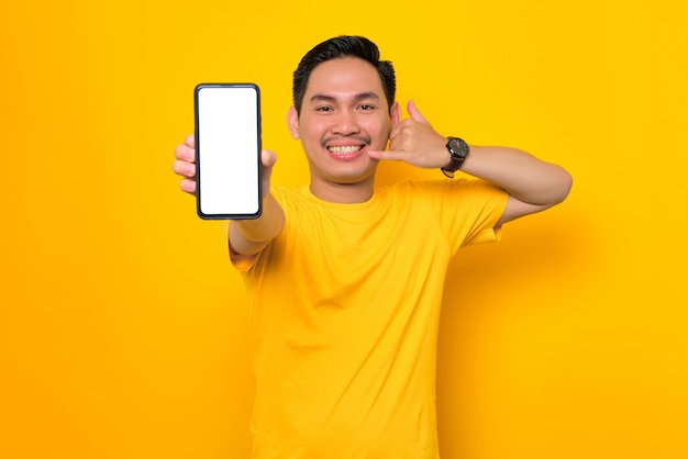 Mockup blank screen cellphone cheerful young asian man in casual tshirt showing mobile phone with blank screen showing call me gesture isolated on yellow background people lifestyle concept