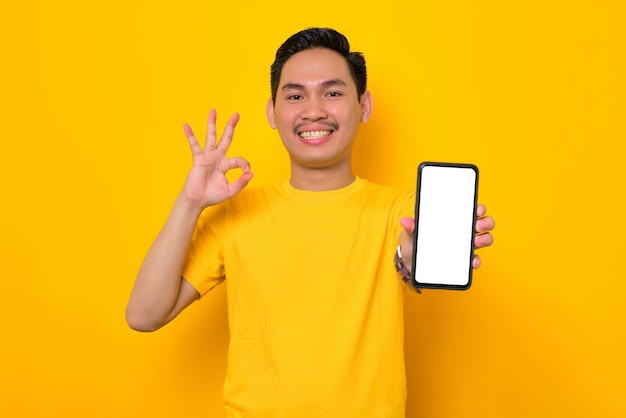 Mockup blank screen cellphone Cheerful young Asian man in casual tshirt showing mobile phone with blank screen showing ok gesture isolated on yellow background People lifestyle concept