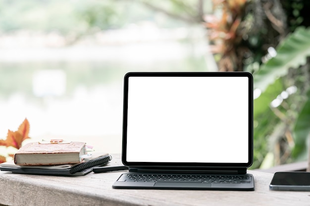 Mockup blank screen tablet with magic keyboard and supplies on the table outdoors, blurred green nature plant on backround.