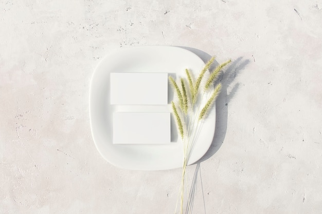 Mockup of business cards on a white plate with dried pampas grass flower on table Flat lay