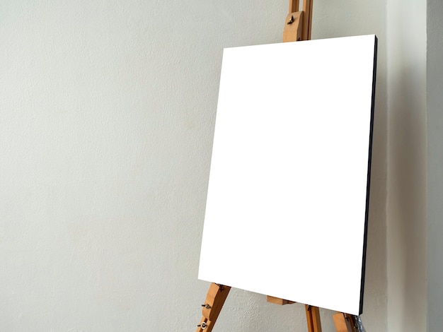 Mockup canvas on easel. White blank art board on wooden easel stand on white wall background in the corner of the artist studio with copy space.