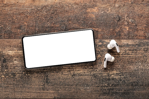 Mockup of cell phone placed horizontally with white blank\
screen and wireless headphones on background of an old wooden table\
game video audio apps music podcasts