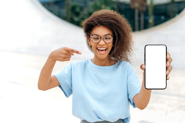 Mockup concept Opgewonden gemengd ras jonge krullende vrouw met bril staat buitenshuis toont smartphone met lege witte mockup scherm wijst vinger ernaar kijkt verbaasd naar camera gelukkig glimlachend