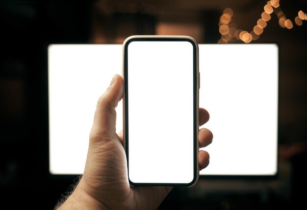 Mockup of hand holding modern smartphone with pc monitor on the background Blank screen