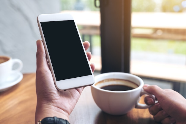 Mockup of hands holding white mobile phone with blank black screen while drinking coffee