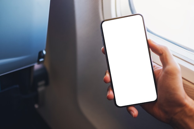 Photo mockup image of a hand holding a black mobile phone with blank desktop screen next to an airplane window