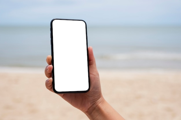 바다 옆에 빈 데스크탑 화면이 있는 검은색 휴대폰을 들고 있는 손의 흉내낸 이미지