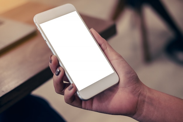 Mockup image of hand holding mobile phone with blank white screen with laptop on wooden table