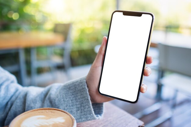 Immagine mockup di una mano che tiene e mostra un telefono cellulare nero con schermo desktop vuoto con tazza di caffè sul tavolo