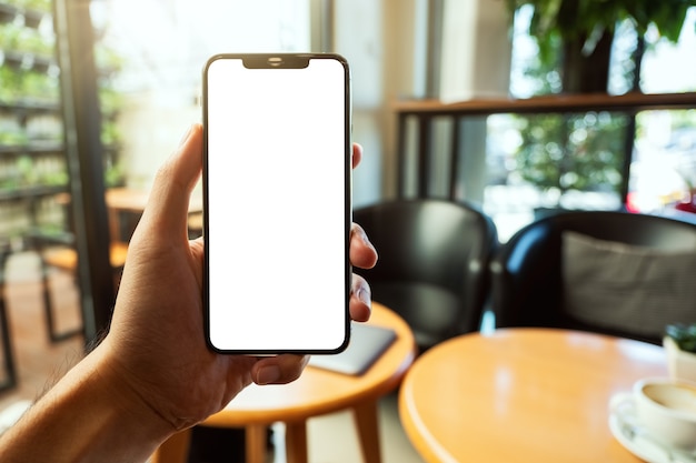 Mockup image of a hand holding and showing black mobile phone with blank white screen in cafe