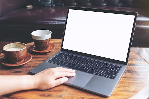 Mockup image of a hand using and typing on laptop with blank white screen and coffee cup on table in modern cafe