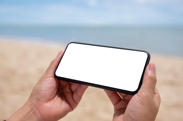 Photo mockup image of hands holding black mobile phone with blank desktop screen by the sea