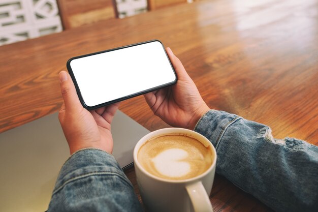 Макет изображения рук, держащих черный мобильный телефон с пустым экраном рабочего стола по горизонтали с ноутбуком и чашкой кофе на столе