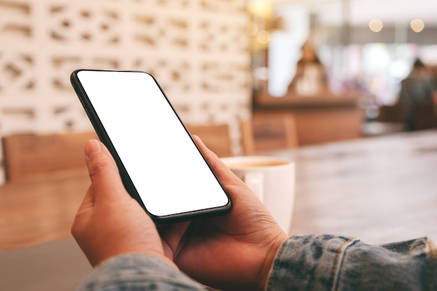 Mockup image of hands holding black mobile phone with blank desktop screen with laptop and coffee cup on the table