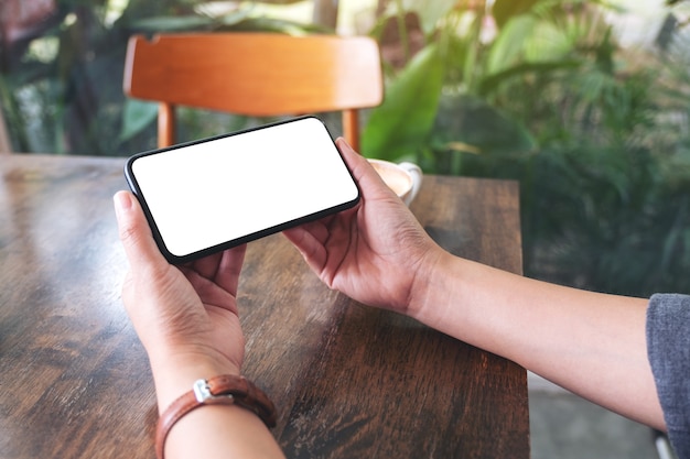 Mockup image of hands holding black mobile phone with blank desktop screen with laptop and coffee cup on the table