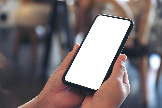 Mockup image of hands holding black mobile phone with blank white desktop screen in cafe