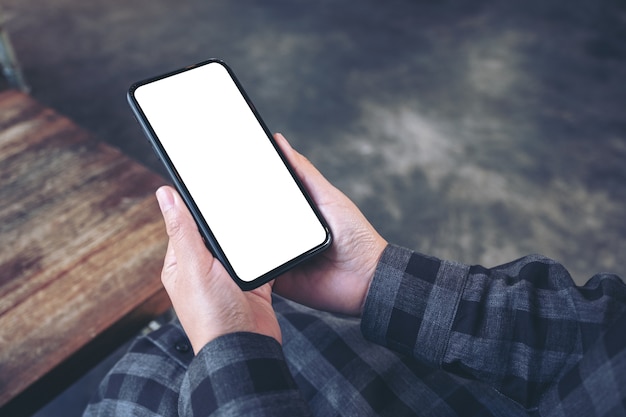 Mockup image of hands holding black mobile phone with blank white screen in vintage cafe