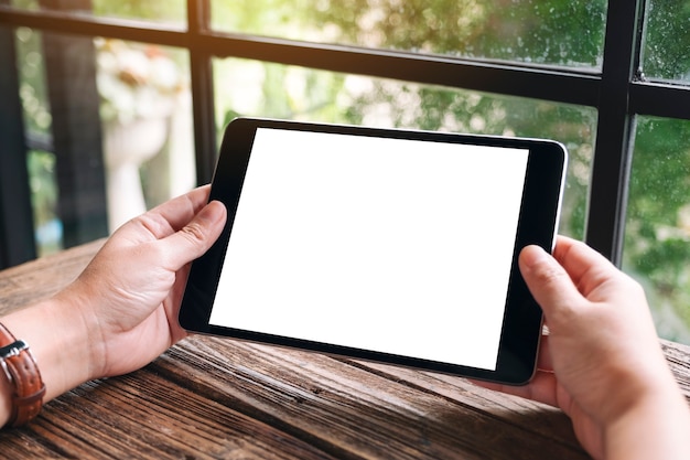 Mockup image of hands holding black tablet pc with blank white desktop screen on wooden table