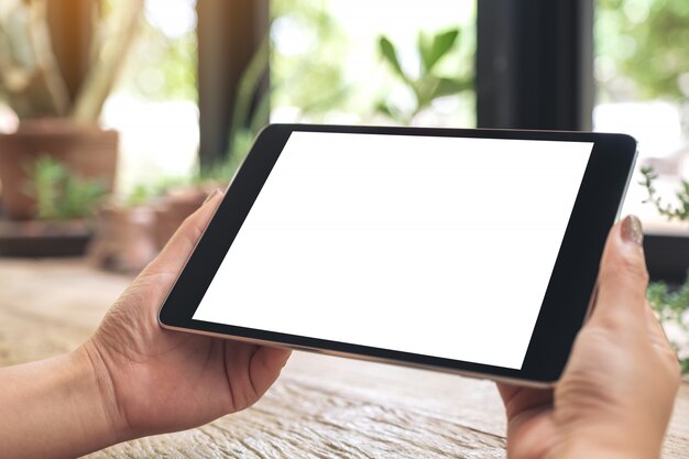 Mockup image of hands holding black tablet pc with blank white desktop screen on wooden table