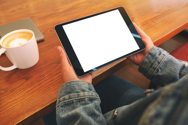 Mockup image of hands holding black tablet pc with blank white screen horizontally with coffee cup on wooden table