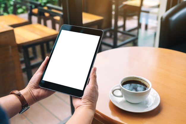 Mockup image of hands holding black tablet pc with blank white screen with coffee cup on wooden table