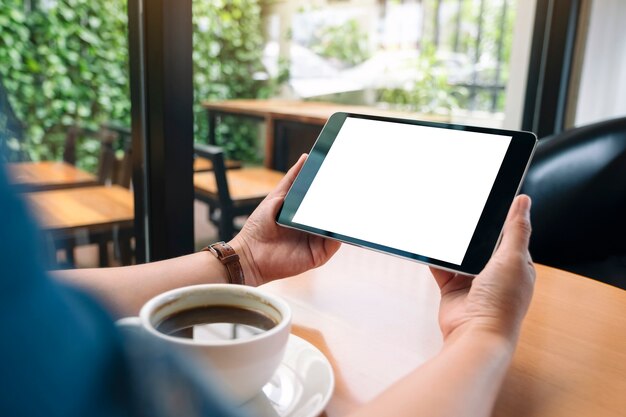 나무 테이블에 커피 컵이 있는 빈 흰색 화면이 있는 검정색 태블릿 PC를 들고 있는 손의 흉내낸 이미지