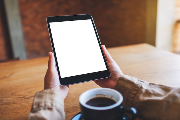 Mockup image of hands holding black tablet pc with blank white screen with coffee cup on wooden table