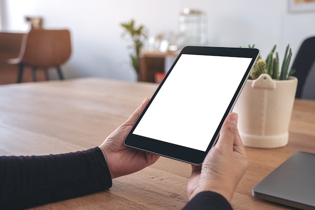 Mockup image of hands holding and using black tablet pc with blank white desktop screen with notebook on wooden table in office