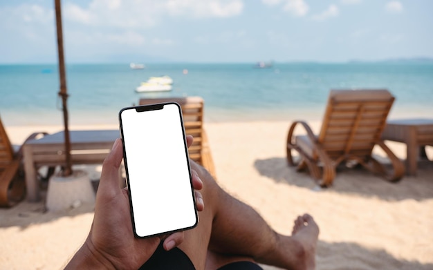 Foto immagine mockup di un uomo che tiene il telefono cellulare con lo schermo del desktop vuoto mentre è seduto sulla spiaggia